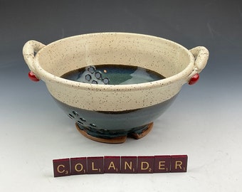 Könnte beinhalten: Ein weißer und schwarzer Keramik-Abtropfkorb mit roten Griffen. Der Abtropfkorb hat eine gesprenkelte Glasur und eine dunkelblaue Glasur auf dem Boden. Das Wort "COLANDER" ist in roten Fliesen ausgeschrieben.