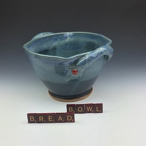Pode incluir: Uma tigela de cerâmica azul com duas alças e detalhes vermelhos. A tigela está sobre uma superfície branca com as palavras "BREAD" e "BOWL" escritas em azulejos vermelhos.