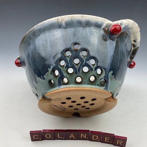 May include: A blue and green ceramic colander with red accents and a white handle. The colander has a decorative design and is handmade. The word "COLANDER" is spelled out in red tiles.