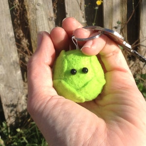 Puede incluir: Un llavero de fieltro verde con forma de fruta pequeña y redonda, con una cara sonriente y dos ojos negros.