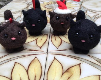 Op de afbeelding: Vier vilten kattenversieringen, twee zwart en twee bruin. Eén bruine kat draagt een rode kerstmanmuts. De versieringen zijn op een betegelde ondergrond met een zonnebloemmotief.