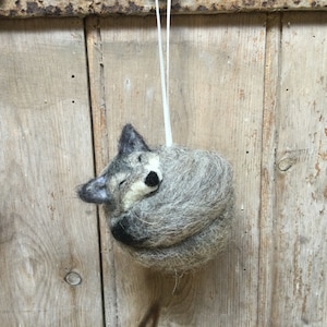 May include: A gray and black felt wolf ornament curled up sleeping. The ornament is hanging from a white string.