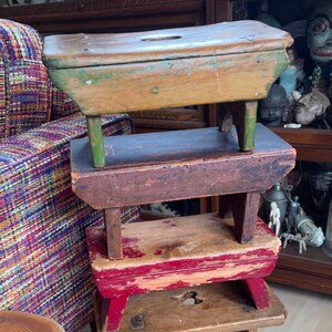 May include: A stack of four wooden stools, each with a different color of paint. The top stool is green, the second is brown, the third is red, and the bottom is natural wood. The stools are stacked in a living room setting.