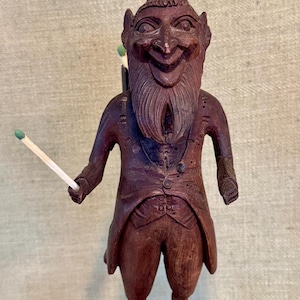 May include: A carved wooden figure of a devil with horns, a beard, and a smiling face. The figure is holding a match in one hand and has matches in a holder on its back. The figure is wearing a suit and stands on a base with several holes.