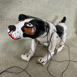 May include: A white, black, and brown wooden puppet dog with black string controls. The dog is posed in a walking position with its head turned to the side.