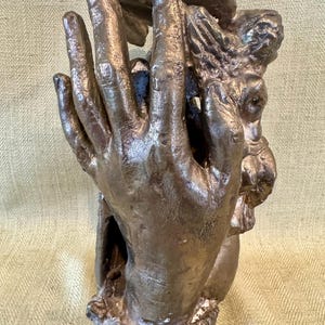 May include: A bronze sculpture of two hands holding a small figure. The sculpture is detailed and has a textured surface.