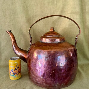 May include: A large, antique copper kettle with a rounded body and a long spout. The kettle has a tarnished finish and a worn handle.