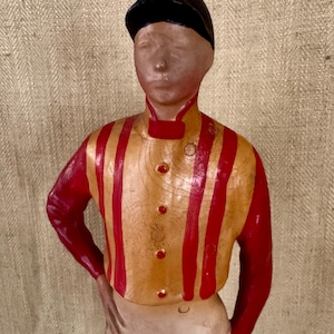 May include: A vintage figurine of a jockey in a racing uniform. The figure wears a black cap, a yellow and red striped shirt, and white trousers. The jockey is posed with one hand on their hip.