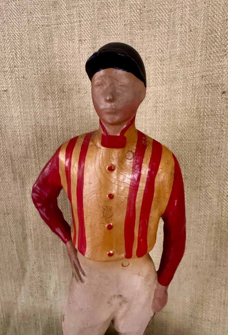 May include: A vintage figurine of a jockey in a racing uniform. The figure wears a black cap, a yellow and red striped shirt, and white trousers. The jockey is posed with one hand on their hip.