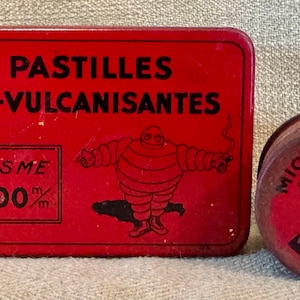 May include: Two vintage red metal containers. One is rectangular and reads "12 PASTILLES AUTO-VULCANISANTES" with a Michelin Man graphic. The other is a round tin with "MICHELIN & CIE" and a Michelin Man graphic.