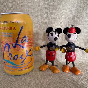 Puede incluir: Una lata de agua con gas La Croix Tangerine junto a dos figuras de madera vintage de Mickey Mouse y Minnie Mouse. La lata es amarilla con texto azul y naranja. Las figuras tienen cuerpos negros, pantalones cortos rojos y manos amarillas.