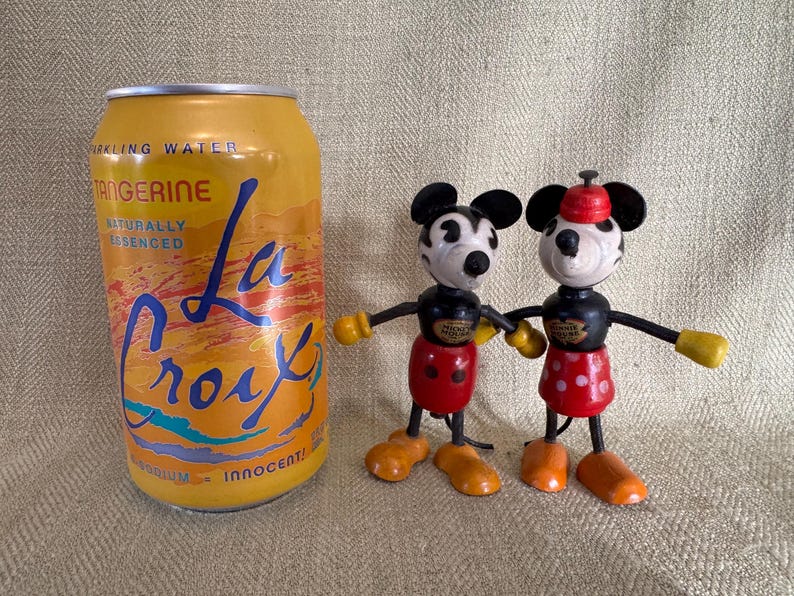 Puede incluir: Una lata de agua con gas La Croix Tangerine junto a dos figuras de madera vintage de Mickey Mouse y Minnie Mouse. La lata es amarilla con texto azul y naranja. Las figuras tienen cuerpos negros, pantalones cortos rojos y manos amarillas.