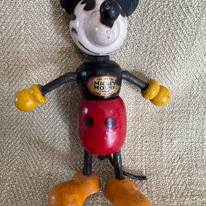 Puede incluir: Juguete de madera vintage de Mickey Mouse con orejas, brazos y piernas negras. La cabeza es blanca con manchas negras, y el cuerpo es rojo y negro. Los brazos tienen guantes amarillos y los pies son naranjas. El juguete tiene una etiqueta que dice "Mickey Mouse".