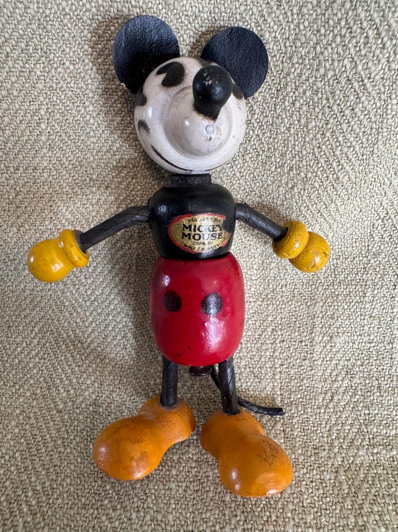 Puede incluir: Juguete de madera vintage de Mickey Mouse con orejas, brazos y piernas negras. La cabeza es blanca con manchas negras, y el cuerpo es rojo y negro. Los brazos tienen guantes amarillos y los pies son naranjas. El juguete tiene una etiqueta que dice "Mickey Mouse".