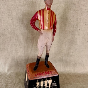 May include: A vintage Jockey underwear advertisement figurine. The figure of a jockey is wearing a red and gold striped shirt, white breeches, black boots, and a black cap. The base is black and brown with the Jockey logo and text.