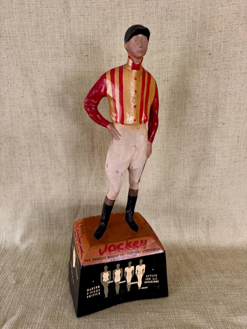 May include: A vintage Jockey underwear advertisement figurine. The figure of a jockey is wearing a red and gold striped shirt, white breeches, black boots, and a black cap. The base is black and brown with the Jockey logo and text.