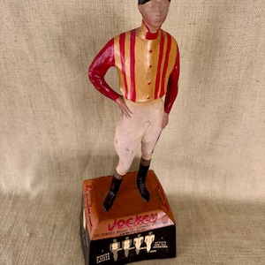 May include: A vintage Jockey figurine of a jockey in a red and gold striped shirt, white breeches, and black boots. The figurine stands on a rectangular base with the word "Jockey" and other text. The jockey is wearing a black cap.