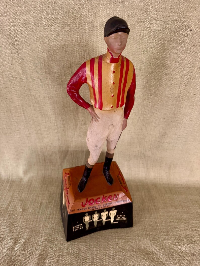 May include: A vintage Jockey figurine of a jockey in a red and gold striped shirt, white breeches, and black boots. The figurine stands on a rectangular base with the word "Jockey" and other text. The jockey is wearing a black cap.