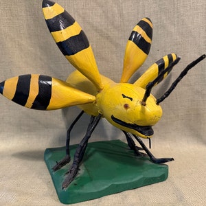 May include: A wooden bee sculpture with yellow and black stripes, painted with a smiling face. The bee is standing on a green base.