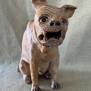 May include: A tan-colored, textured ceramic sculpture of a seated bulldog with a wrinkled face, prominent lower teeth, and a small round bell attached to a collar around its neck. The figure has dark, expressive eyes and pointed ears.