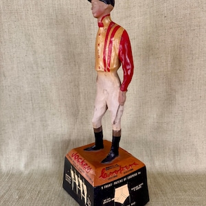 May include: A vintage wooden jockey figurine, wearing a black cap, red and gold jacket, white breeches, and black boots. The figurine stands on a black and orange base with text that reads "Jockey" and "Coopers".