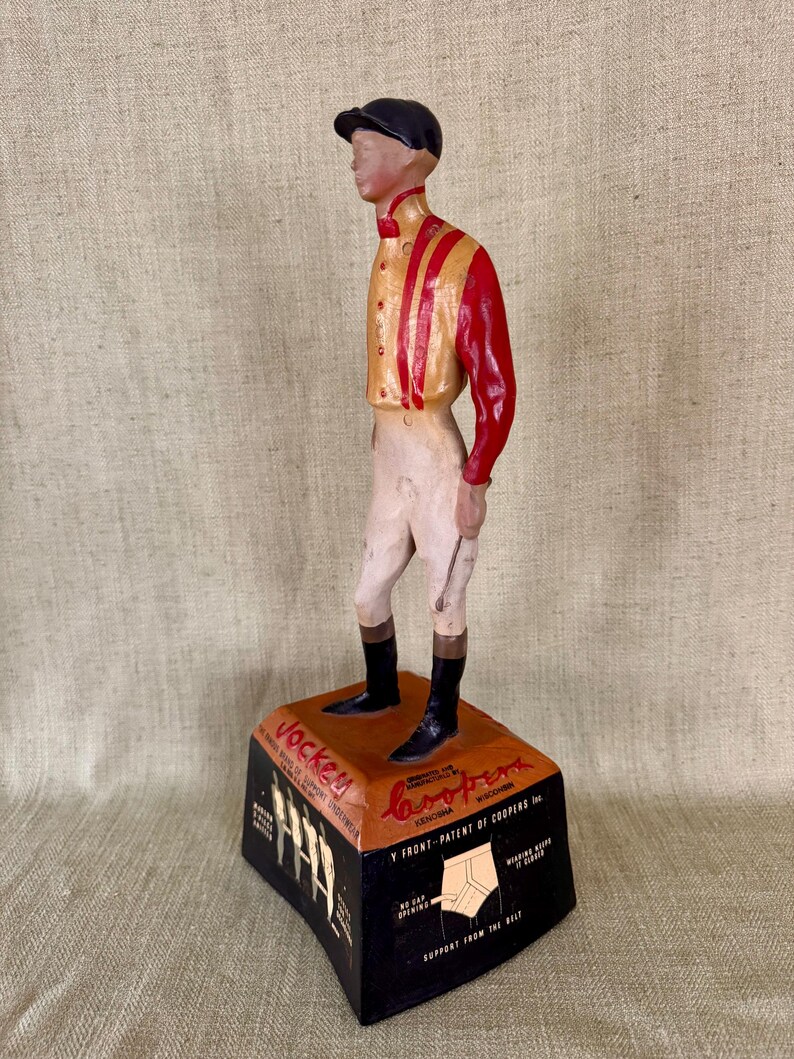 May include: A vintage wooden jockey figurine, wearing a black cap, red and gold jacket, white breeches, and black boots. The figurine stands on a black and orange base with text that reads "Jockey" and "Coopers".