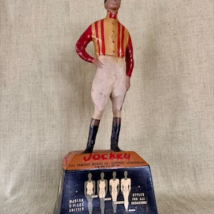 May include: A vintage Jockey underwear advertisement featuring a figurine of a jockey in a red and yellow striped shirt, white breeches, and black boots. The base displays the Jockey logo and text: "THE FAMOUS BRAND OF SUPPORT UNDERWEAR".