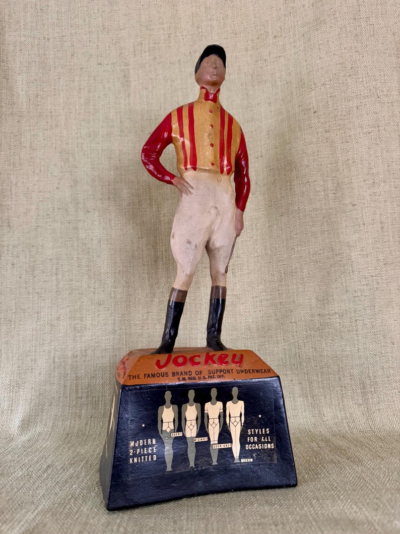 May include: A vintage Jockey underwear advertisement featuring a figurine of a jockey in a red and yellow striped shirt, white breeches, and black boots. The base displays the Jockey logo and text: "THE FAMOUS BRAND OF SUPPORT UNDERWEAR".