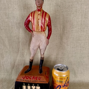 May include: A vintage Jockey underwear display with a jockey figurine in a red and yellow striped shirt, white breeches, and black boots. The base displays the Jockey logo and product information. A can of La Croix Tangerine sparkling water is next to the display.