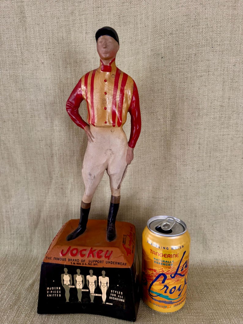 May include: A vintage Jockey underwear display with a jockey figurine in a red and yellow striped shirt, white breeches, and black boots. The base displays the Jockey logo and product information. A can of La Croix Tangerine sparkling water is next to the display.