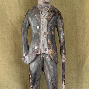 May include: A weathered wooden sculpture of a figure wearing a top hat and a suit, standing on a circular base. The figure has a distressed grey and white finish, with visible wood grain and paint wear, holding a thin cane in the left hand.