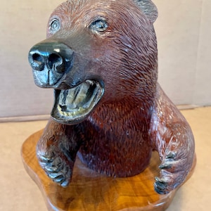 May include: A carved wooden bear head sculpture with a toothy grin, set on a wooden base. The bear's fur is detailed with brown and black paint.