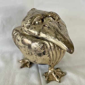 May include: A silver-colored pelican figurine with a detailed face and webbed feet. The pelican's beak is open and the figurine is standing on a white surface.