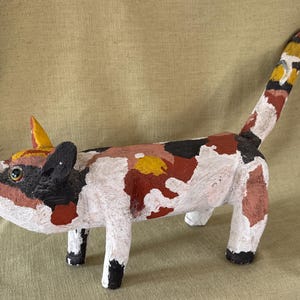 May include: A handcrafted cat figurine featuring a textured, multi-colored surface with patches of white, black, rust-red, and yellow. The cat stands on four legs with a long, upright tail, pointed ears, and dark eyes, showcasing a painted, uneven finish.