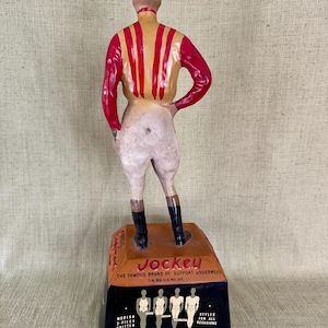 May include: A vintage Jockey underwear advertisement figurine. The jockey figure wears a red and yellow striped shirt, white breeches, and black boots. The base displays the Jockey logo and brand information.