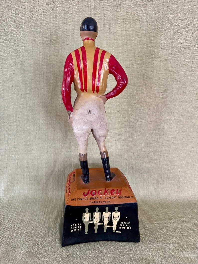 May include: A vintage Jockey underwear advertisement figurine. The jockey figure wears a red and yellow striped shirt, white breeches, and black boots. The base displays the Jockey logo and brand information.
