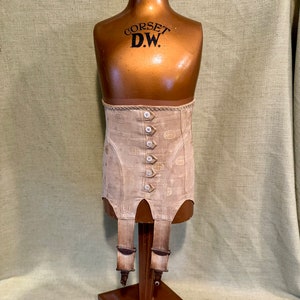 May include: A vintage beige corset with a decorative pattern and button closures. The corset is displayed on a wooden mannequin with the text "CORSET D.W." on the chest. The corset has four suspender straps attached to the bottom.