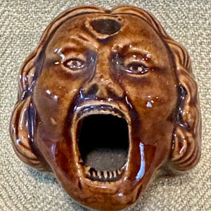 May include: A brown ceramic ashtray shaped like a screaming face. The ashtray has a wide open mouth and a hole on top of the head. The face has detailed features, including eyes, eyebrows, and wavy hair. The ashtray is glazed and has a glossy finish.