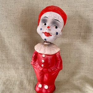 May include: A vintage toy clown with a red and white costume. The clown has a painted face with black eyes, a red mouth, and blue tear drops. The clown is made of paper mache and has a spring-loaded mechanism in its body.