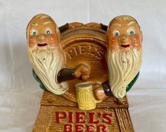May include: A ceramic piggy bank shaped like two gnomes sitting on a barrel with a beer mug. The barrel is labeled "Piel's" and the base says "Piel's Beer".