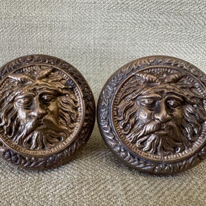 May include: Two antique bronze cabinet knobs. Each knob features a detailed relief of a bearded face within a circular frame. The knobs have a weathered, aged appearance, suggesting they are vintage or vintage-inspired.