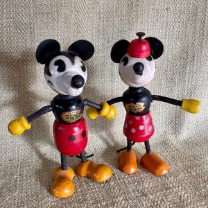 Puede incluir: Dos figuras de madera vintage de Mickey y Minnie Mouse. Mickey tiene cabeza y orejas negras, cara blanca, pantalones cortos rojos y guantes y zapatos amarillos. Minnie lleva una falda roja con lunares blancos y un sombrero rojo.