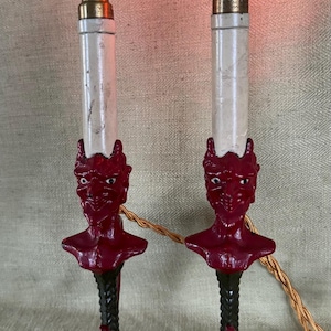 May include: Pair of vintage devil-themed electric candle lamps. Each lamp features a red devil face base, a white candle, and a flame-shaped light bulb. The lamps have a black and red claw-foot base and a gold-colored cord.