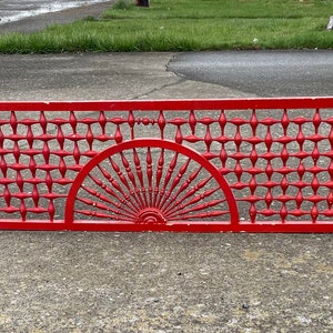 May include: A red metal fence panel with a decorative design featuring a sunburst pattern in the center and a grid pattern on either side.