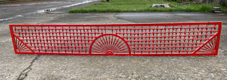 May include: A red metal fence panel with a decorative design featuring a sunburst pattern in the center and a grid pattern on either side.