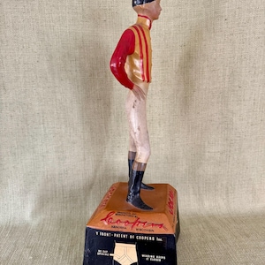 May include: A vintage figurine of a jockey in a red and gold uniform, black boots, and a black cap. The figurine stands on a rectangular base with the text "Coopers Kenosha Wisconsin" and "Y FRONT-PATENT OF COOPERS Inc."