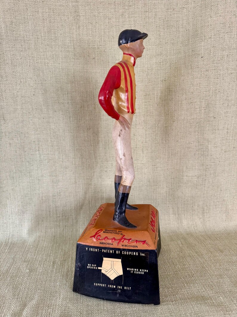 May include: A vintage figurine of a jockey in a red and gold uniform, black boots, and a black cap. The figurine stands on a rectangular base with the text "Coopers Kenosha Wisconsin" and "Y FRONT-PATENT OF COOPERS Inc."