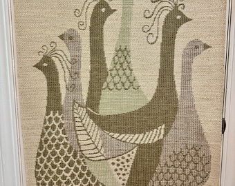 May include: A woven wall hanging featuring a stylized design of three birds with long necks and intricate patterns. The birds are in shades of brown, green, and purple, and the background is a light beige.