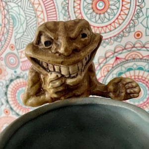 May include: A ceramic figurine of a grinning, grotesque creature with a wide mouth and large teeth. The creature is holding a mug with a dark gray interior.