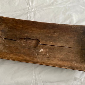 May include: A long, wooden dough bowl with a natural brown finish. The bowl has a slightly curved shape and shows signs of age and use, including a crack down the center.